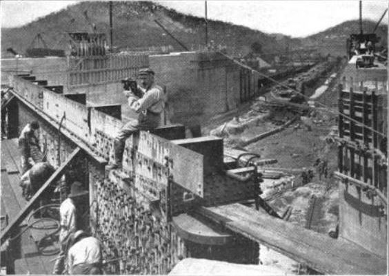 A man sits on top of one of the gates of the Panama Canal locks under construction. He is holding a camera.